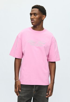 Νέος άνδρας που φοράει ένα ροζ T-shirt Wood Wood Field Studies και σκούρο τζιν, στέκεται μπροστά σε ένα μονόχρωμο ανοιχτό φόντο, κοιτώντας προς τα αριστερά του.