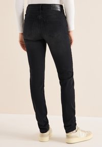 Jeans de denim negro de corte ajustado con una cintura de altura media, que presentan un brillo sutil, bolsillos estándar y una cinturilla con marca. Calzado visible abajo.