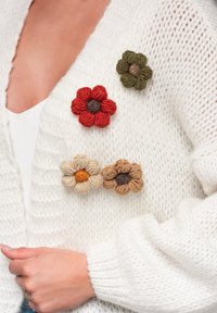 Vit stickad kofta med fyra blommig broscher i tyg: röd, grön, beige och brun, där varje blomma har texturerade kronblad och kontrasterande mitt.