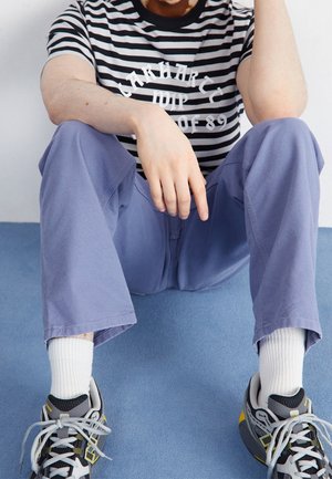 Camiseta a rayas en blanco y negro con gráfico blanco, combinada con pantalones morados claros y zapatillas grises con detalles amarillos y calcetines blancos.