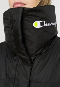 Schwarze Daunenjacke mit hohem Kragen, ausgestattet mit einem weißen und neon-gelben "Champion"-Logo. Glatte, glänzende Textur mit gesteppten Paneelen.