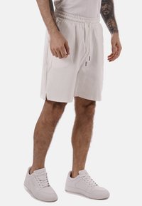 Pantaloni corti bianchi con vita elastica, stringa regolabile e spacco laterale. Indossati con sneaker bianche, caratterizzate da una texture liscia.