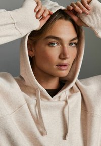 Sweat à capuche beige clair avec une finition texturée, doté d'une capuche réglable et de poignets côtelés, porté par-dessus un haut marron.