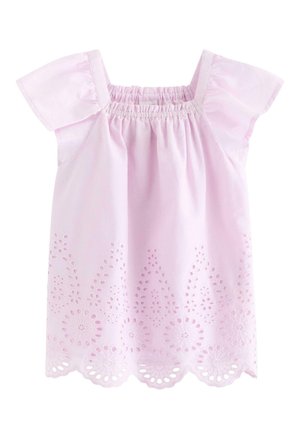 Smilende ung pige iført matchende lyse lilla top og bukser med hulmønster, solhat og mørke flettede sko, stående foran en ensfarvet baggrund.