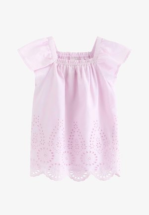 Uśmiechnięta młoda dziewczyna w pasującym jasnofioletowym komplecie z ażurowej tkaniny, kapeluszu przeciwsłonecznym i ciemnych plecionych butach, stojąca na tle jednolitego tła.