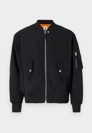 Sort bomberjakke med frontlynlås, grå T-shirt, lyseblå vide jeans, hvide sneakers og en sort kasket med tekst.