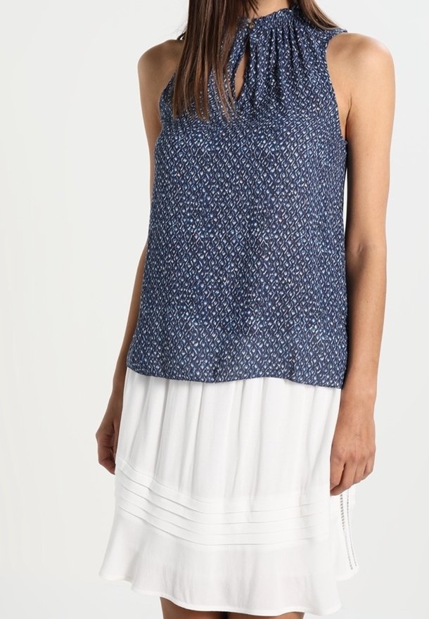 Femme portant un haut bleu marine à motifs sans manches avec une encolure en trou de serrure et une jupe plissée blanche longueur genou.