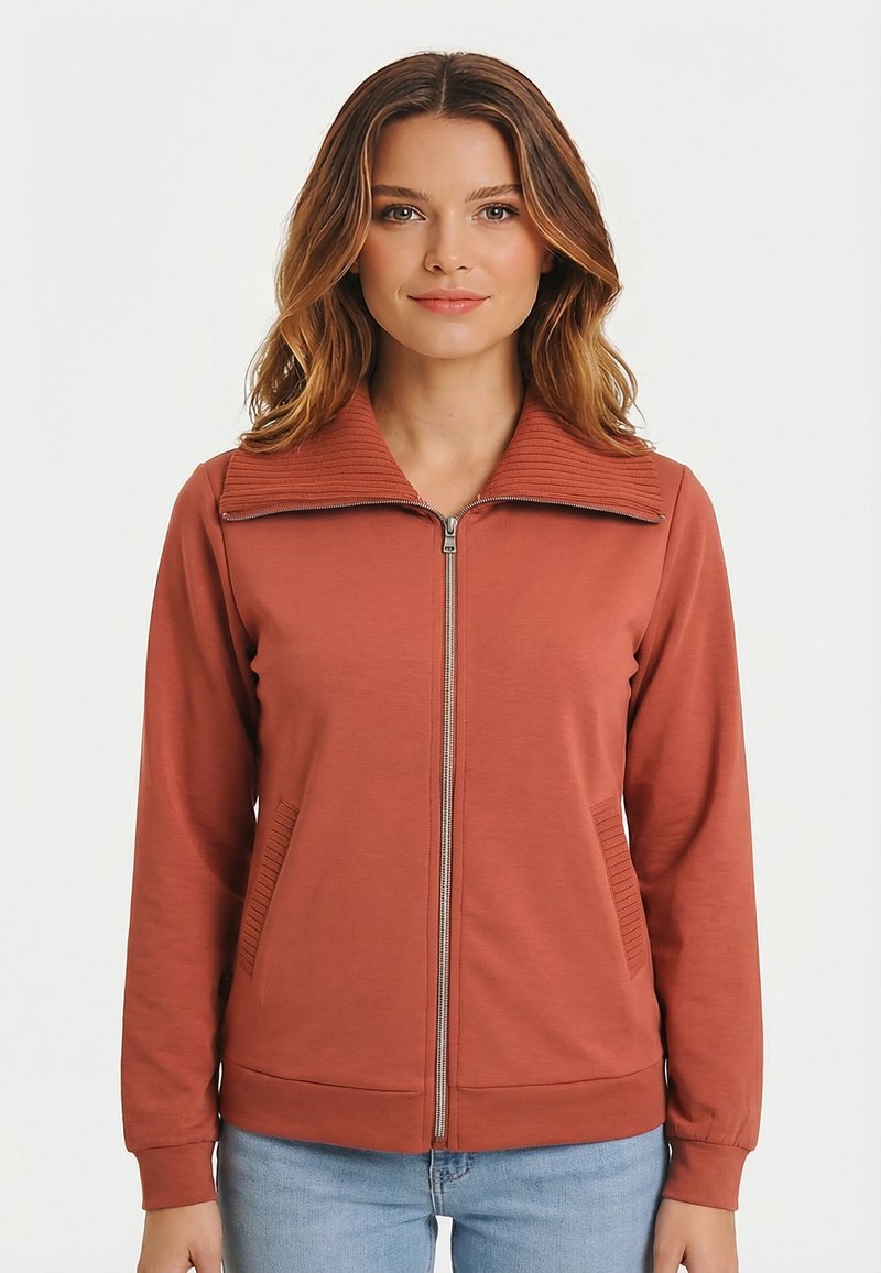 Mujer con chaqueta con cremallera de color óxido, con cuello acanalado y detalles en los bolsillos, combinada con jeans azul claro, mirando hacia el frente.