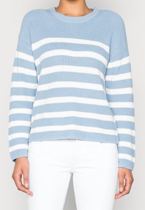 Personne portant un pull en tricot à rayures horizontales bleu clair et blanc avec un pantalon blanc, debout devant un fond uni.