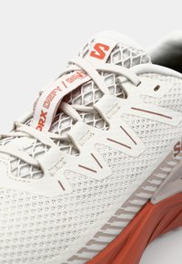 Hvit atletisk sko med mesh-overdel, røde detaljer og en teksturert såle. Har et distinkt snøringdesign og merkevare på tungen.