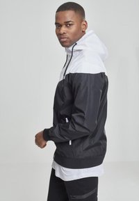 Chaqueta cortavientos en blanco y negro con capucha, que presenta un ajuste relajado, puños elásticos y bolsillos laterales con cremallera. Textura de nylon suave.