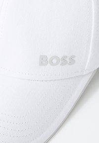 Hvid kasket med en struktureret design, der har en tekstureret stofoverflade og et sølvlogo "BOSS" på forsiden.