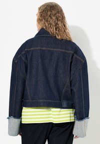 Denimjack met een relaxte pasvorm, donkerblauwe kleur, contrasterend stiksel en brede grijze manchetten. Gelaagd met een gestreept geel en wit shirt eronder.