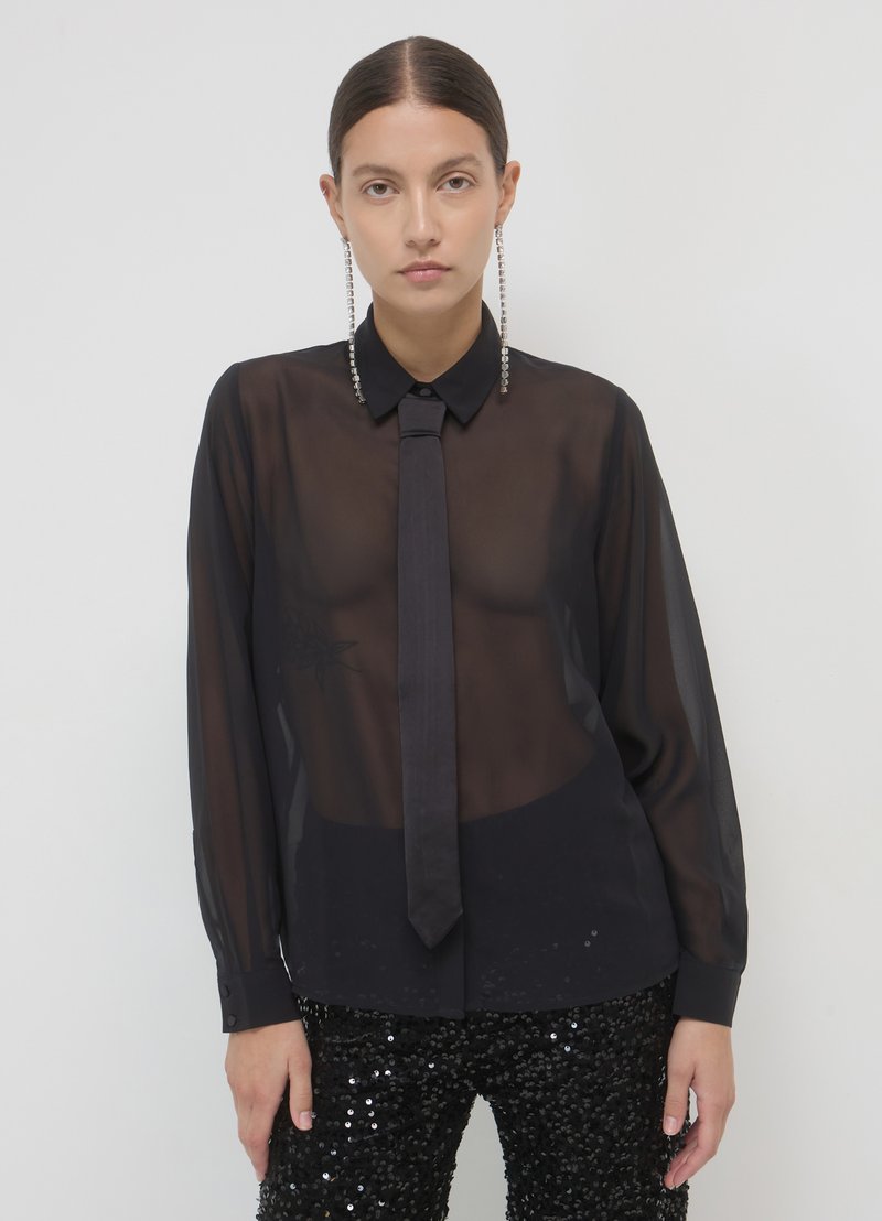 Blouse noire transparente avec un col classique, une cravate assortie et des manches longues. Associée à un pantalon noir à sequins pour un contraste texturé.