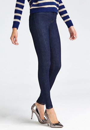 Personne portant un jean skinny bleu foncé, un pull rayé bleu et blanc, et des chaussures à talons hauts métalliques à bouts pointus, debout avec les jambes croisées.