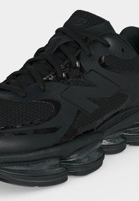 Chaussure de sport noire avec une semelle épaisse comportant des coussinets visibles, une tige en mesh et synthétique, et une fermeture à lacets.
