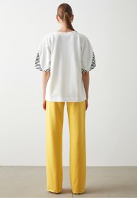 Bílé oversized tričko s černými pruhovanými krátkými rukávy, doplněné vysokými žlutými kalhotami rovného střihu. Hladká tkanina, minimalistický design.