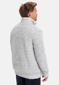 Mann, der nach rechts schaut, trägt einen dicken, grauen Strickpullover mit hohem Kragen und dunklen Hosen vor einem weißen Hintergrund.