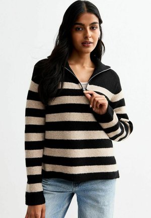 Pull rayé noir et beige avec une demi-fermeture éclair, texture côtelée, manches longues et coupe décontractée. Porté avec un jeans bleu clair.