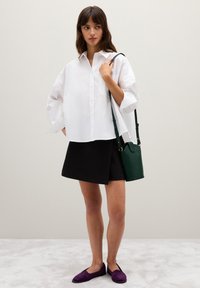 Camicia bianca oversize con bottoni, gonna nera avvolgente, scarpe slip-on viola e borsa a tracolla in pelle verde. Texture morbide con linee semplici.