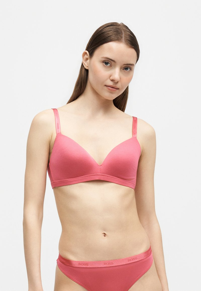 Jeune femme aux longs cheveux bruns portant un ensemble soutien-gorge et sous-vêtements rose assorti, debout devant un fond blanc uni.