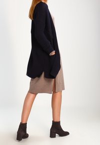 Marinblå stickad cardigan med öppen front och långa ärmar, som bärs tillsammans med en beige åtsittande klänning och mörka ankelstövlar, vilket visar textur och en avslappnad silhuett.