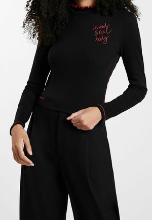 Nainen pukeutuneena mustaan pitkähihainen paita, jossa on punaiset tikkauskoristeet ja rinnassa kirjailtu teksti ”mind soul body”, yhdistettynä mustiin leveälahkeisiin housuihin.