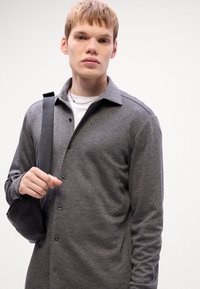 Grå skjorta med krage, långa ärmar och en mjuk textur. Modellen bär en liten svart väska över axeln.