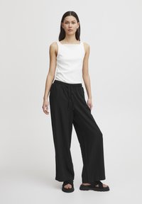 Balts bez piedurknēm tops ar kvadrātveida dekoltē, apvienots ar brīviem melniem biksēm un melniem sandālēm; mīksts audums, brīva piegriezuma, vienkāršs dizains.