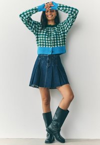 Vrouw die glimlacht met opgestoken armen, gekleed in een groen-blauw houndstooth vest, een geplooide denim minirok en kniehoge zwarte laarzen tegen een witte muur.