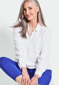 Weißes Bluse mit langen Ärmeln, kombiniert mit blauen knöchellangen Hosen. Die Accessoires umfassen mehrere Ringe und ein silbernes Armband.
