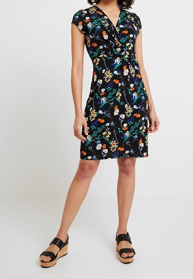 Femme portant une robe noire à manches courtes avec un imprimé floral coloré et des sandales compensées noires à semelles en liège, debout sur un fond uni.