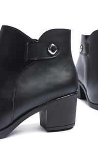 Botas de tobillo de cuero negro con punta redondeada, borde superior con ondas y detalle de botón de metal brillante. Tacón cuadrado con una superficie texturizada.