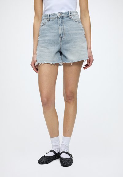 Personne portant un short en jean bleu clair taille haute avec des ourlets effilochés, des chaussettes blanches et des chaussures Mary Jane noires, debout devant un fond uni.