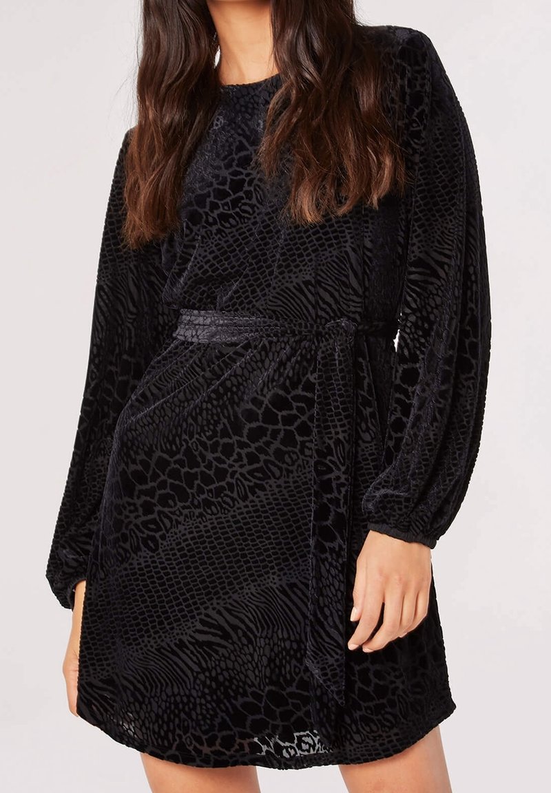 Femme portant une robe noire à manches longues avec un motif animal texturé et une ceinture en tissu assortie nouée à la taille.
