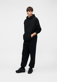 Μαύρο φούτερ με κουκούλα, τσέπη τύπου πουγκί και κορδόνι στη κουκούλα, σε συνδυασμό με μαύρο παντελόνι χαλαρής γραμμής. Τα παπούτσια έχουν χοντρές λεπτομέρειες και υφή.