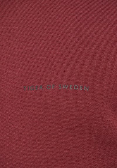 Bordo adīts audums ar centrētu "Tiger of Sweden" logo, kas izdrukāts mazos, mazos, tumši pelēkos burtos.
