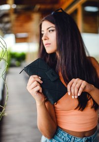Black quilted wallet with a textured surface, featuring a flap and zipper closure, held by a person standing in a wooden outdoor setting.
