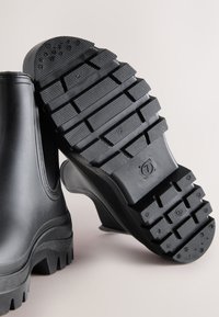 Pair of black rubber boots with chunky, textured soles shown against a plain light background.