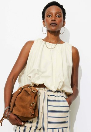 Femme aux cheveux courts porte un haut blanc sans manches, un pantalon rayé, de grandes boucles d'oreilles et tient un sac à main en suède marron sur fond blanc.