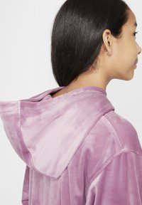 Kapuca, plišasta vijolična jakna z mehko teksturo, z okroglo kapuco in vidnimi šivi. Preprost dizajn brez okrasnih elementov.