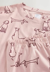 Pehmeässä vaaleanpunaisessa puuvillapukeutumisessa on toistuva burgundinvärinen koira-kuvitus. Settiin kuuluu lyhythihainen paita ja elastisella vajaamittaisella helmalla varustetut shortsit.