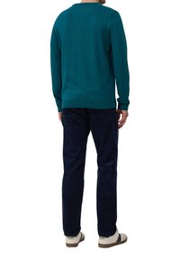 Mann in einem tealfarbenen Langarmpullover, dunklen blauen Hosen und weißen Turnschuhen, steht mit dem Rücken zu einem weißen Hintergrund.