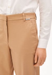 Beige broek met een gladde textuur, voorzien van een rechte snit en een zijzak. Label zichtbaar op de tailleband; gedragen met een wit shirt.