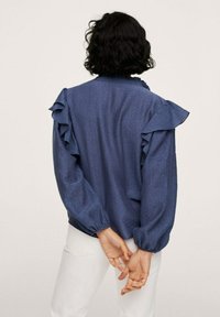 Blue long-sleeve blouse with ruffled shoulder details, textured fabric, and elastic cuffs. Worn with white pants.