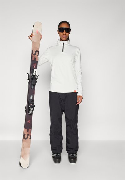 Χειμερινός εξοπλισμός: μια λευκή φανέλα με φερμουάρ, μαύρα παντελόνια σκι και ένα ζευγάρι σκούρα γυαλιά ηλίου. Ο σκιέρ κρατά ένα καφέ σκι με μαύρες βάσεις.
