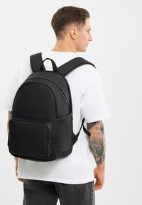 Fekete hátizsák sima szintetikus anyagból, elülső zsebbel és párnázott vállpántokkal, egy fehér inget viselő személyen bemutatva.