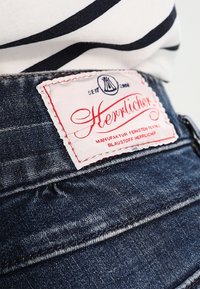 Farkkukankaiset shortsit tummansinisellä pesulla, joissa on voimakas valkoinen ja punainen etiketti, jossa lukee "Herrlicher". Ompeleet näkyvissä.