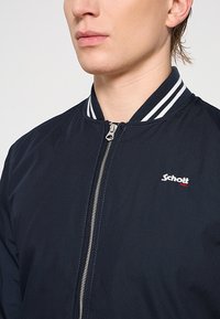 Chaqueta bomber azul marino con diseño de cierre frontal, cuello de canalé blanco y azul marino, y un pequeño logotipo blanco y rojo en el lado izquierdo del pecho.