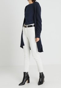 Marinblå ribbade koftan över en marinblå topp, i kombination med vita jeans och svarta ankellånga stövlar. Den har en lång design med öppen framsida och figursydda ärmar.